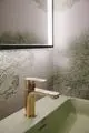 Goldene Waschtischarmatur von Ideal Standard mit minimalistischer Form und leichtem Zugang zum Wasserhahn. Hintergründe mit grünen, dekorativen Mustern.